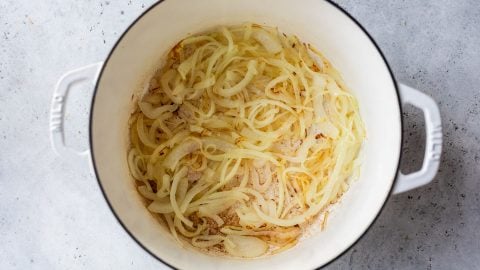 Thinly sliced onions cooking and softening in a large white Dutch oven on a light gray surface.