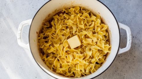 A white pot filled with cooked egg noodles and a pat of butter on top, sitting on a light gray surface.