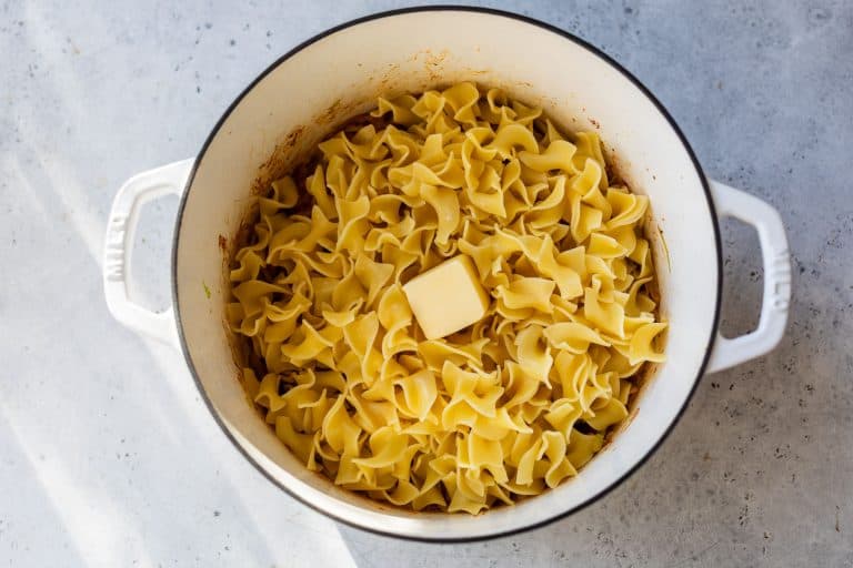A white pot filled with cooked egg noodles and a pat of butter on top, sitting on a light gray surface.