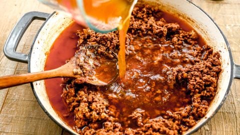 Ground beef simmering in a pan as broth is poured in, with a wooden spoon stirring the mixture.