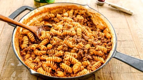 A skillet filled with rotini pasta, ground beef, and tomato sauce, being stirred with a wooden spoon on a wooden table.