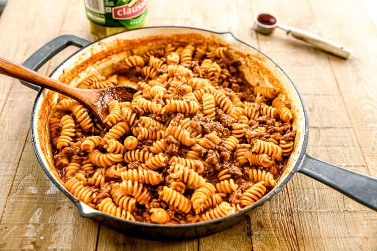 A skillet filled with rotini pasta, ground beef, and tomato sauce, being stirred with a wooden spoon on a wooden table.