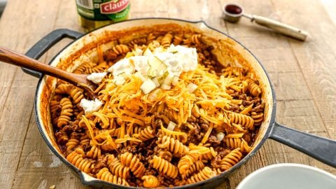 A skillet of rotini pasta with ground meat, shredded cheddar, chopped onions, and sour cream on top, on a wooden table.