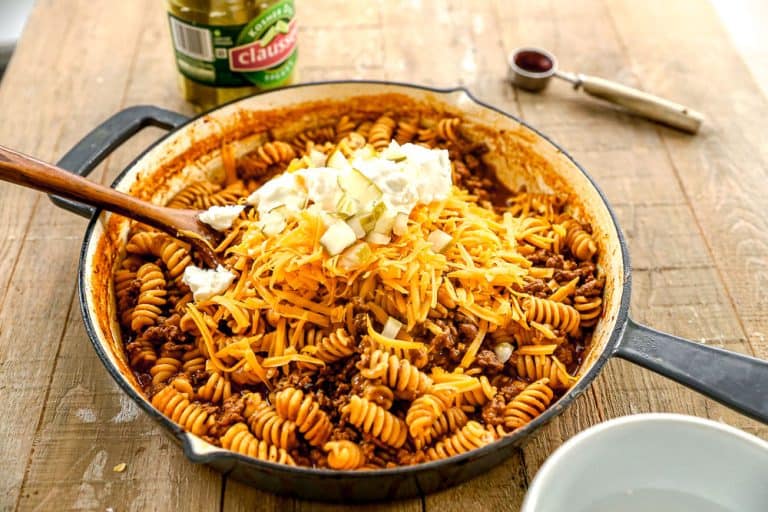 A skillet of rotini pasta with ground meat, shredded cheddar, chopped onions, and sour cream on top, on a wooden table.