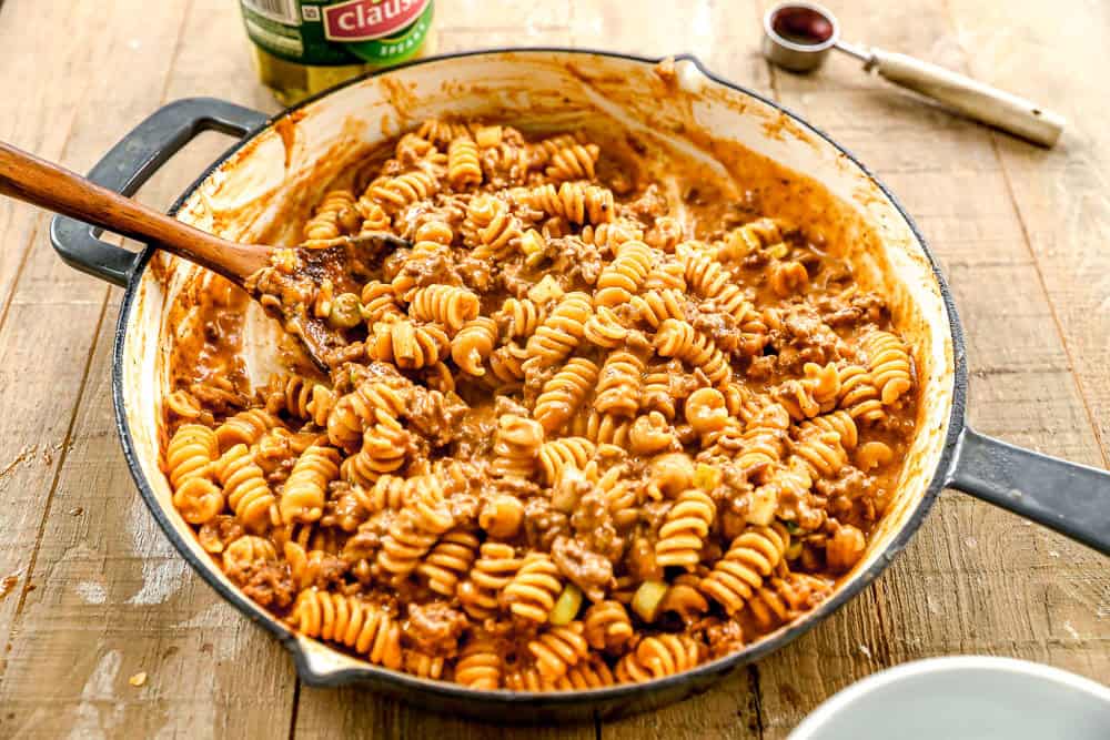 A skillet filled with cheeseburger casserole and ground meat sauce, with a wooden spoon for stirring.
