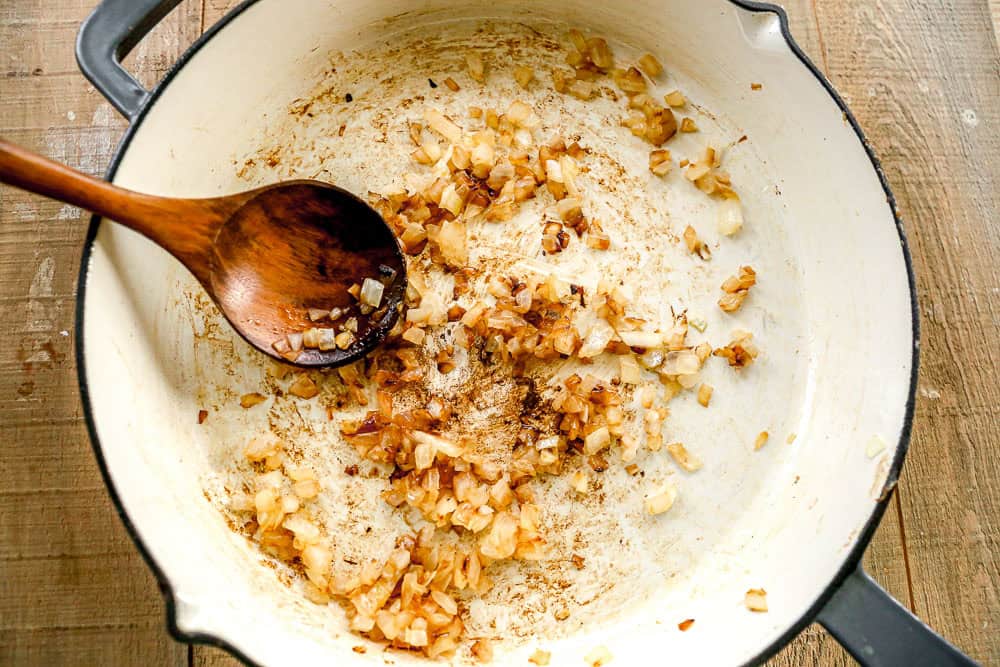 Chopped onions sautรฉing and browning in a white pot with a wooden spoon on a wooden surface.
