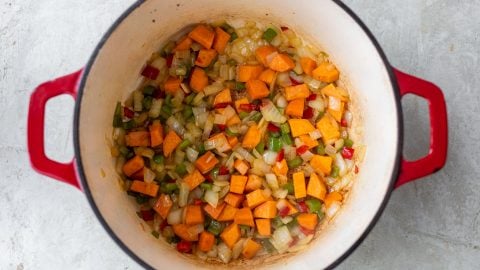 Diced sweet potatoes, onions, and bell peppers sautéing in a red Dutch oven on a light surface.