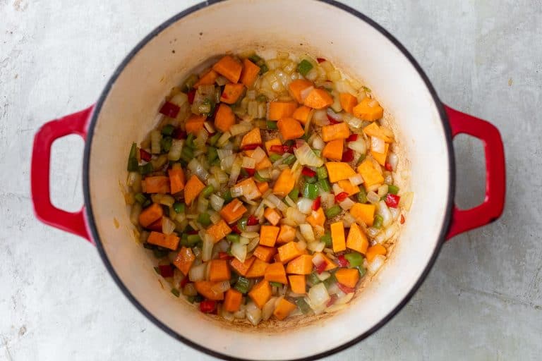 Diced sweet potatoes, onions, and bell peppers sautéing in a red Dutch oven on a light surface.