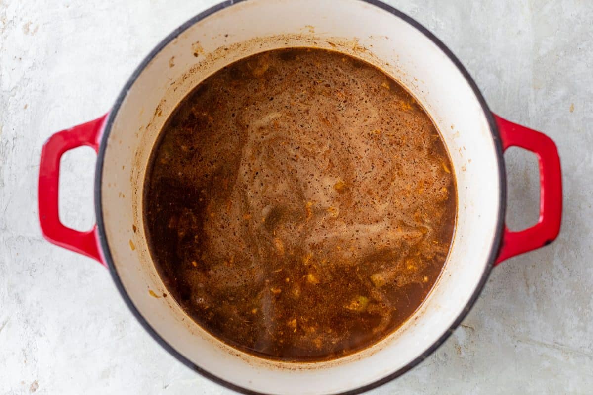 A red Dutch oven filled with simmering dark brown broth sits on a light gray surface.