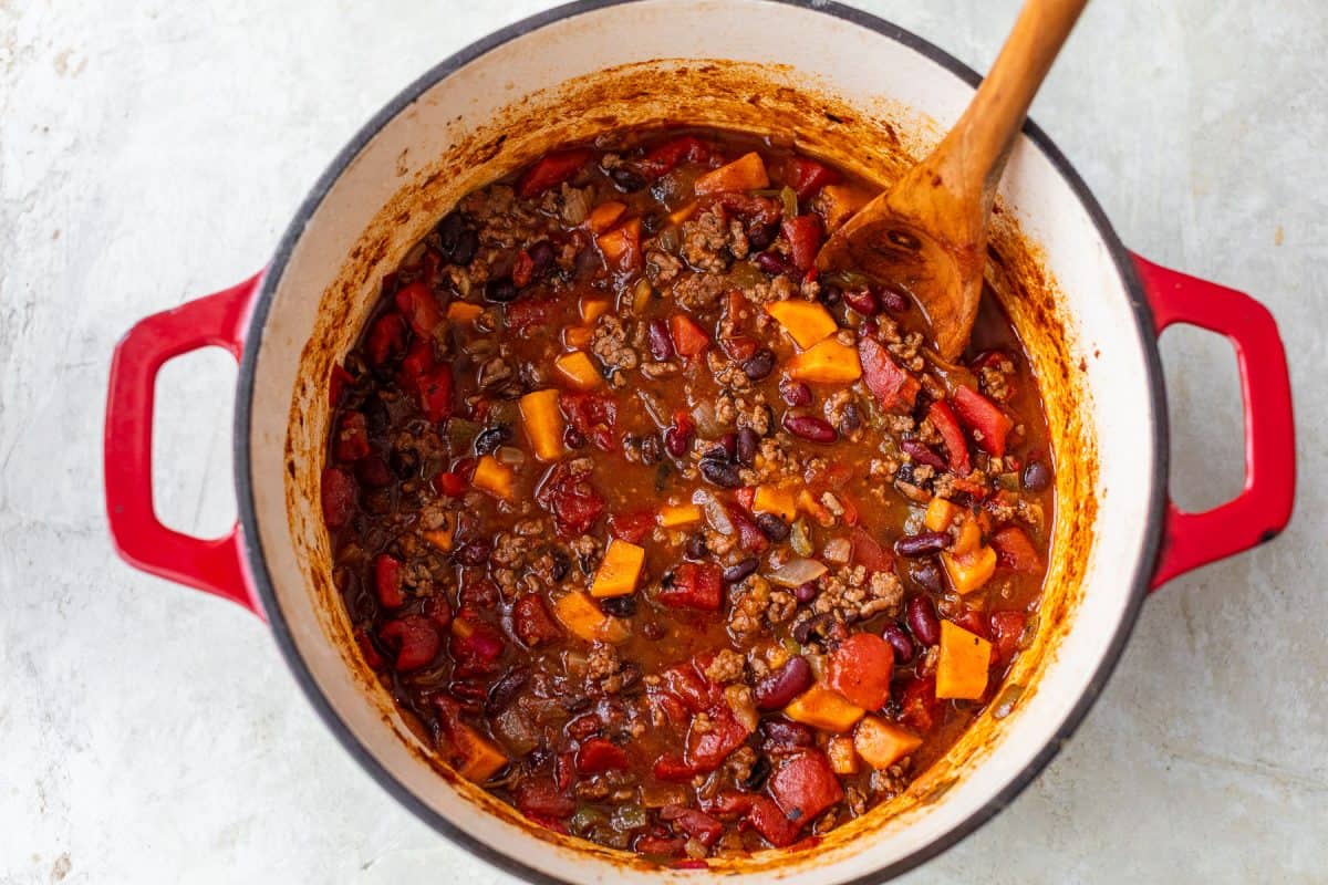 A pot of chili with beans, tomatoes, ground beef, and sweet potatoes, stirred with a wooden spoon.