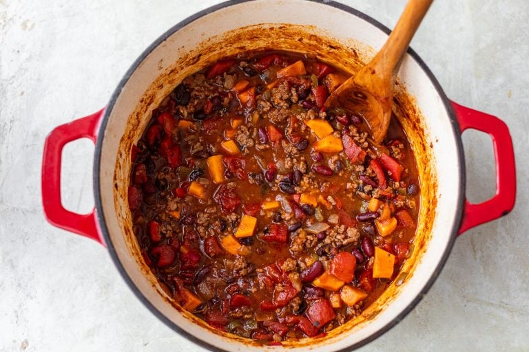 A pot of chili with beans, tomatoes, ground beef, and sweet potatoes, stirred with a wooden spoon.