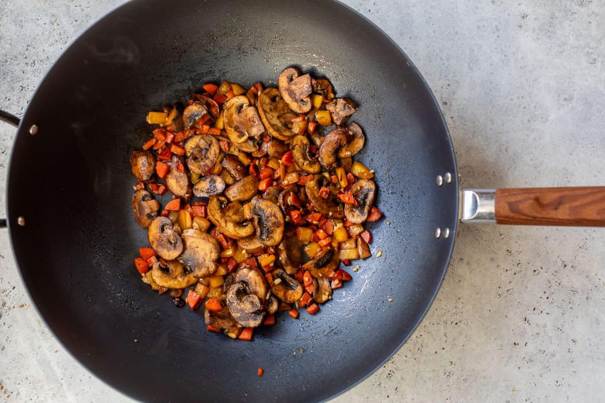 Sliced mushrooms and diced vegetables are being stir-fried in a black wok on a light-colored countertop.