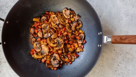Sliced mushrooms and diced vegetables are being stir-fried in a black wok on a light-colored countertop.