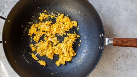 Scrambled eggs cooking in a black nonstick pan with a wooden handle on a light gray countertop.
