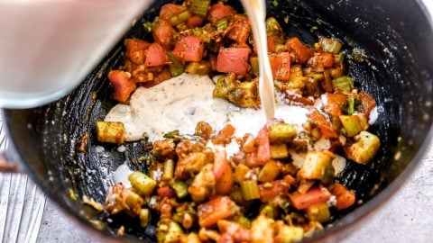 Milk is being poured into a pan with chopped vegetables, including tomatoes, celery, and spices, for cooking.