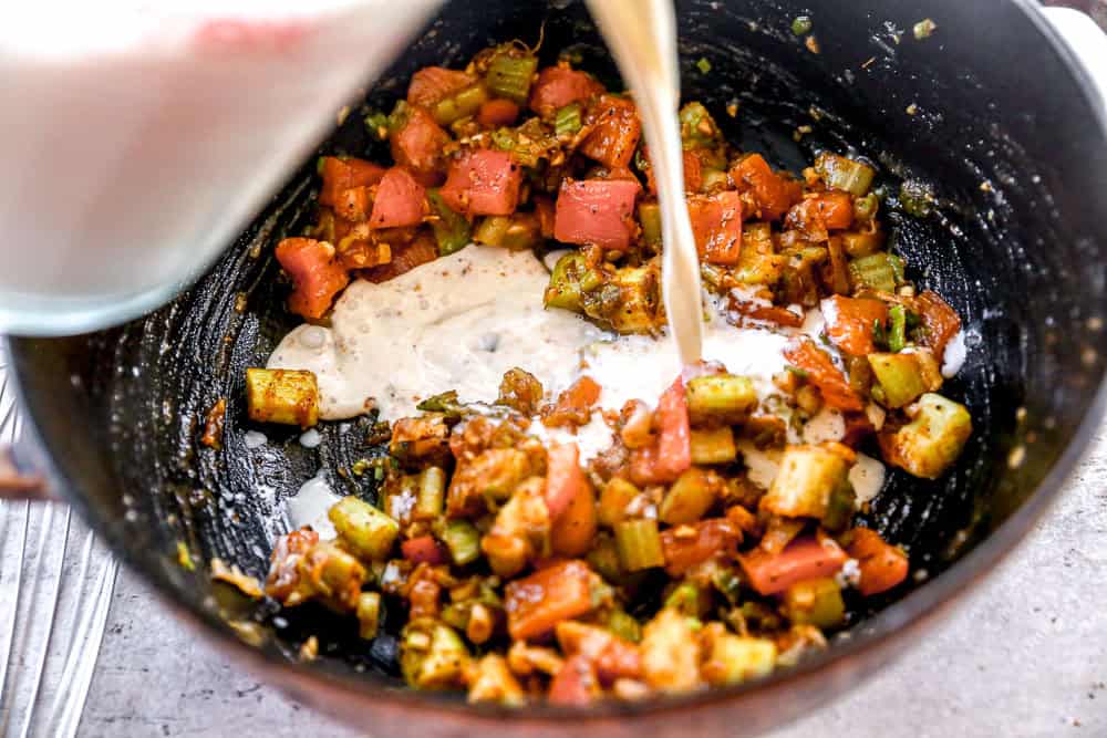 Milk is being poured into a pan with chopped vegetables, including tomatoes, celery, and spices, for cooking.