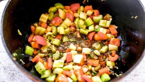Chopped celery, red peppers, and green onions sautรฉing in a pot with herbs and oil.