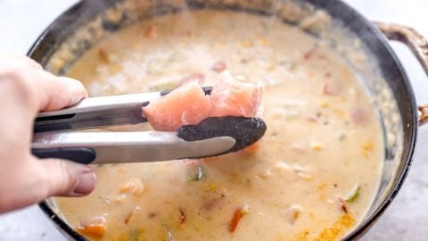 A hand holds tongs with raw salmon over a pot of creamy chowder with vegetables.
