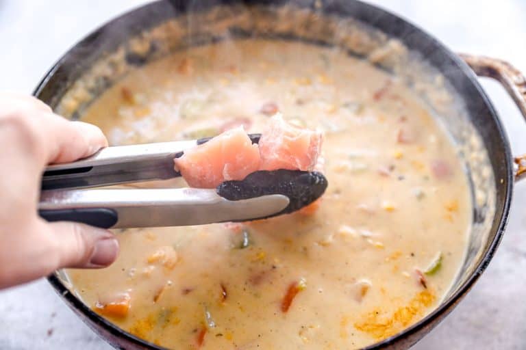 A hand holds tongs with raw salmon over a pot of creamy chowder with vegetables.