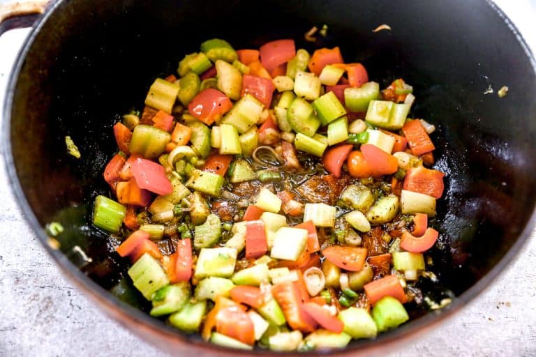 Chopped celery, red peppers, and green onions sautéing in a pot with herbs and oil.