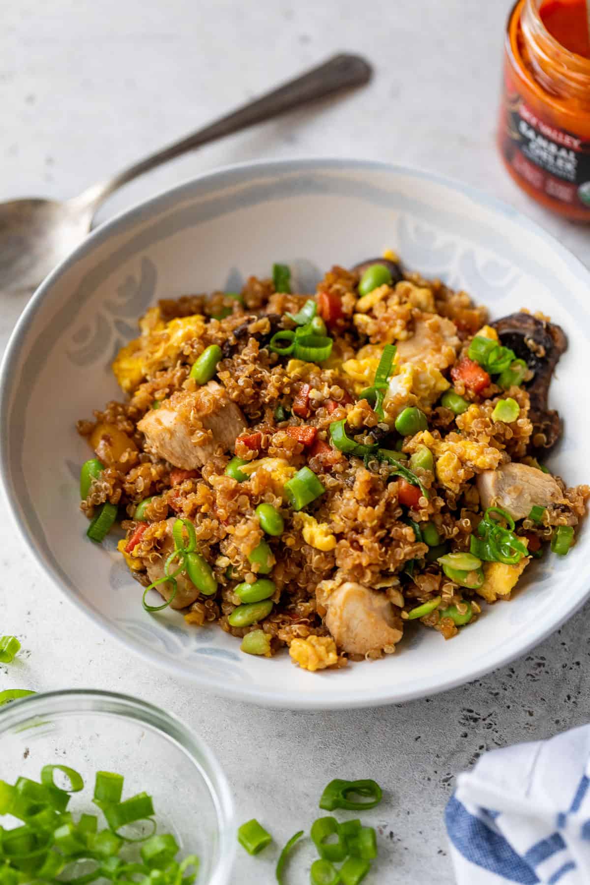 A bowl of quinoa fried rice with chicken, vegetables, and green onions, with a spoon and green onions nearby.