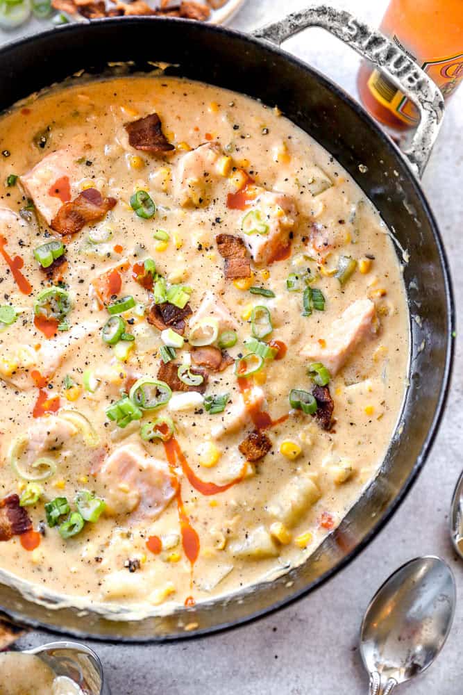 A pot of creamy salmon chowder with corn, potatoes, bacon, green onions, and a drizzle of hot sauce.