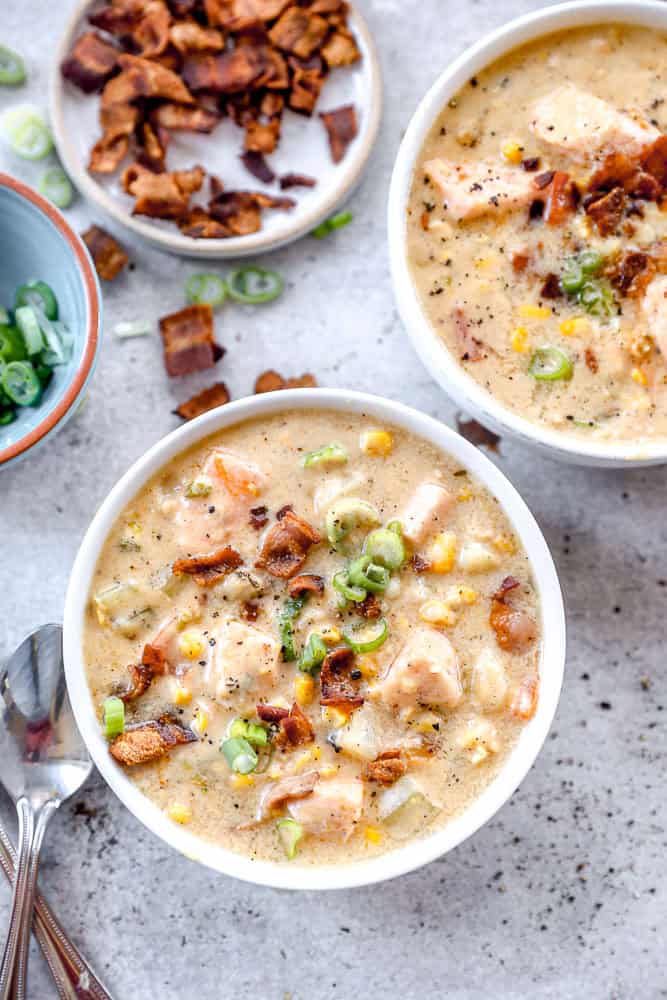 Two bowls of creamy salmon chowder soup with corn, bacon pieces, and chopped green onions, with extra toppings on the side.