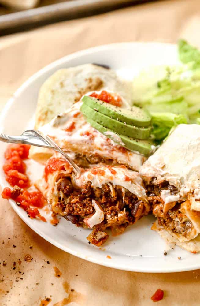 A fork cuts into a cheesy beef burrito topped with avocado, served with salsa, lettuce, and bread on a white plate.