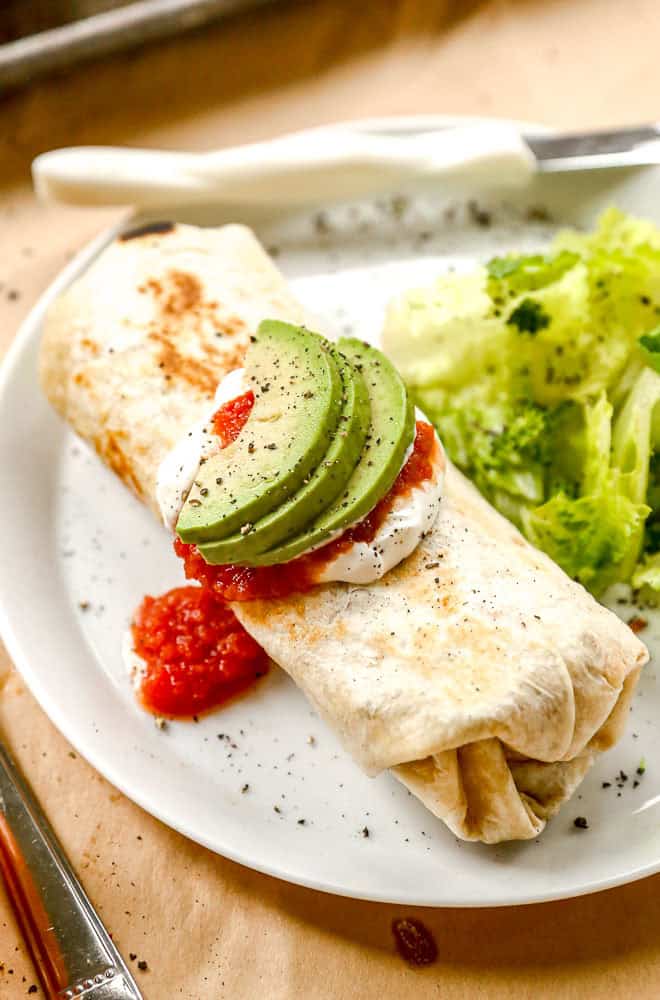 A beef burrito topped with avocado slices, sour cream, and salsa, served with lettuce on a white plate.