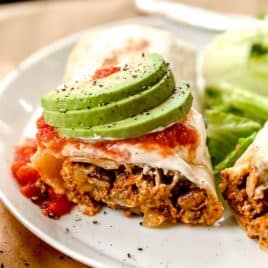 A burrito topped with salsa and sliced avocado, served with lettuce on a white plate.