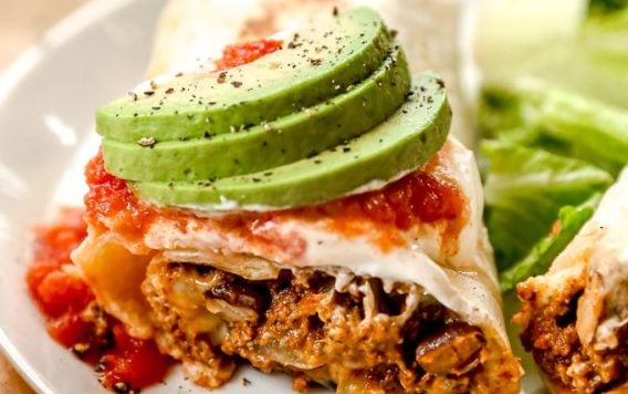 A burrito topped with salsa and sliced avocado, served with lettuce on a white plate.
