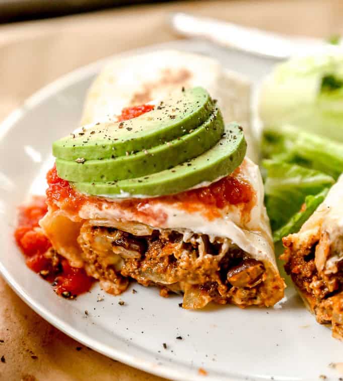 A ground beef burrito topped with salsa and sliced avocado, served with lettuce on a white plate.