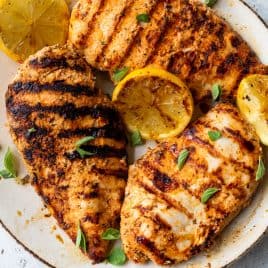 Grilled chicken breasts with char marks, garnished with lemon slices and fresh herbs on a ceramic plate.
