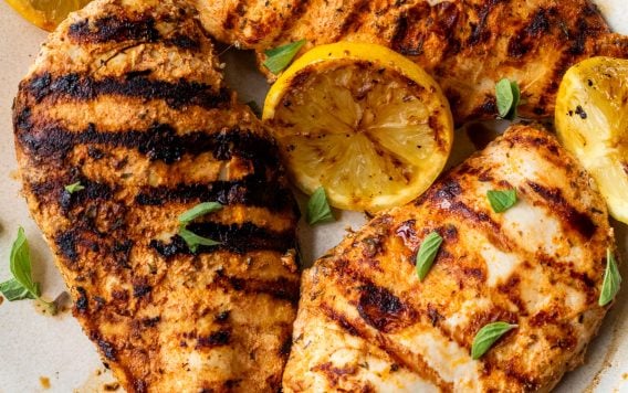 Grilled chicken breasts with char marks, garnished with lemon slices and fresh herbs on a ceramic plate.