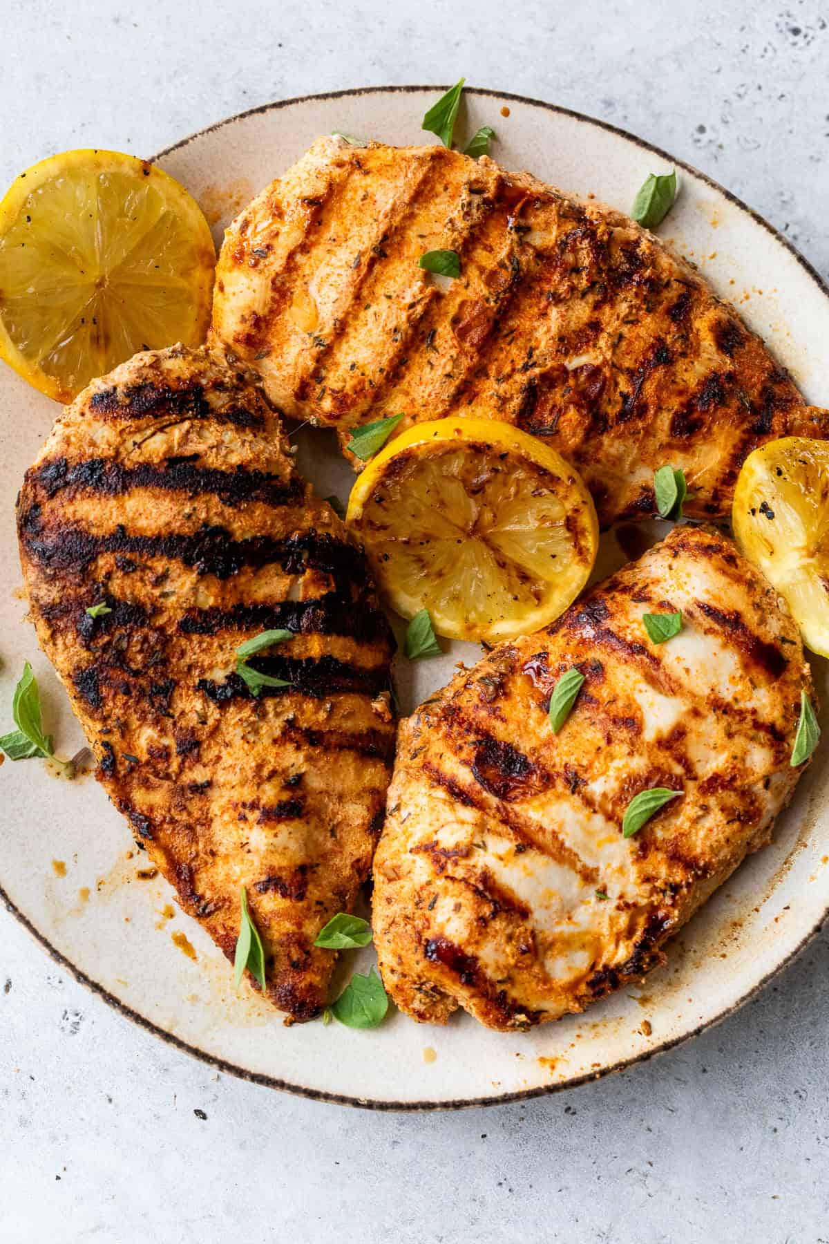 Grilled chicken breasts with char marks, garnished with lemon slices and fresh herbs on a ceramic plate.