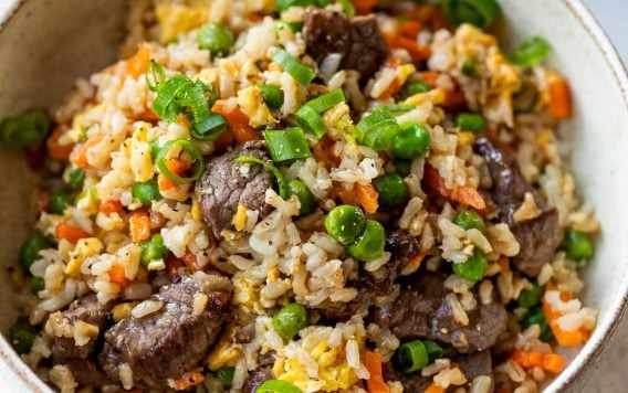 A bowl of steak fried rice with beef, peas, carrots, scrambled egg, and chopped green onions.