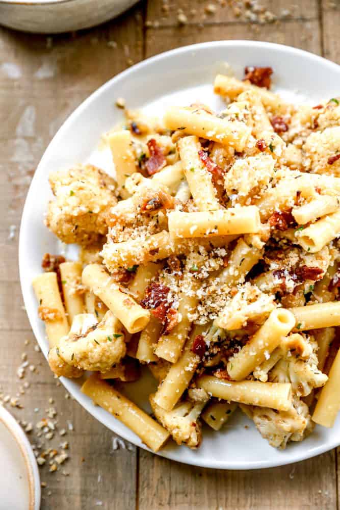 Cauliflower pasta with bacon and breadcrumbs on a white plate, set on a rustic wooden table.