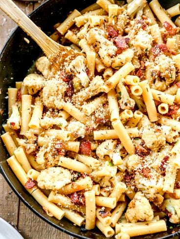 Ziti pasta with roasted cauliflower, bacon, and breadcrumbs in a skillet, mixed with a wooden spoon.