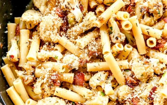 Ziti pasta with roasted cauliflower, bacon, and breadcrumbs in a skillet, mixed with a wooden spoon.