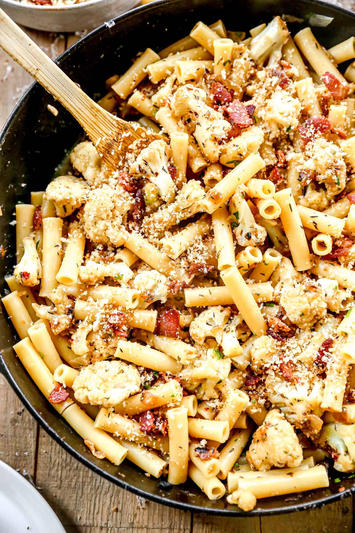 Ziti pasta with roasted cauliflower, bacon, and breadcrumbs in a skillet, mixed with a wooden spoon.