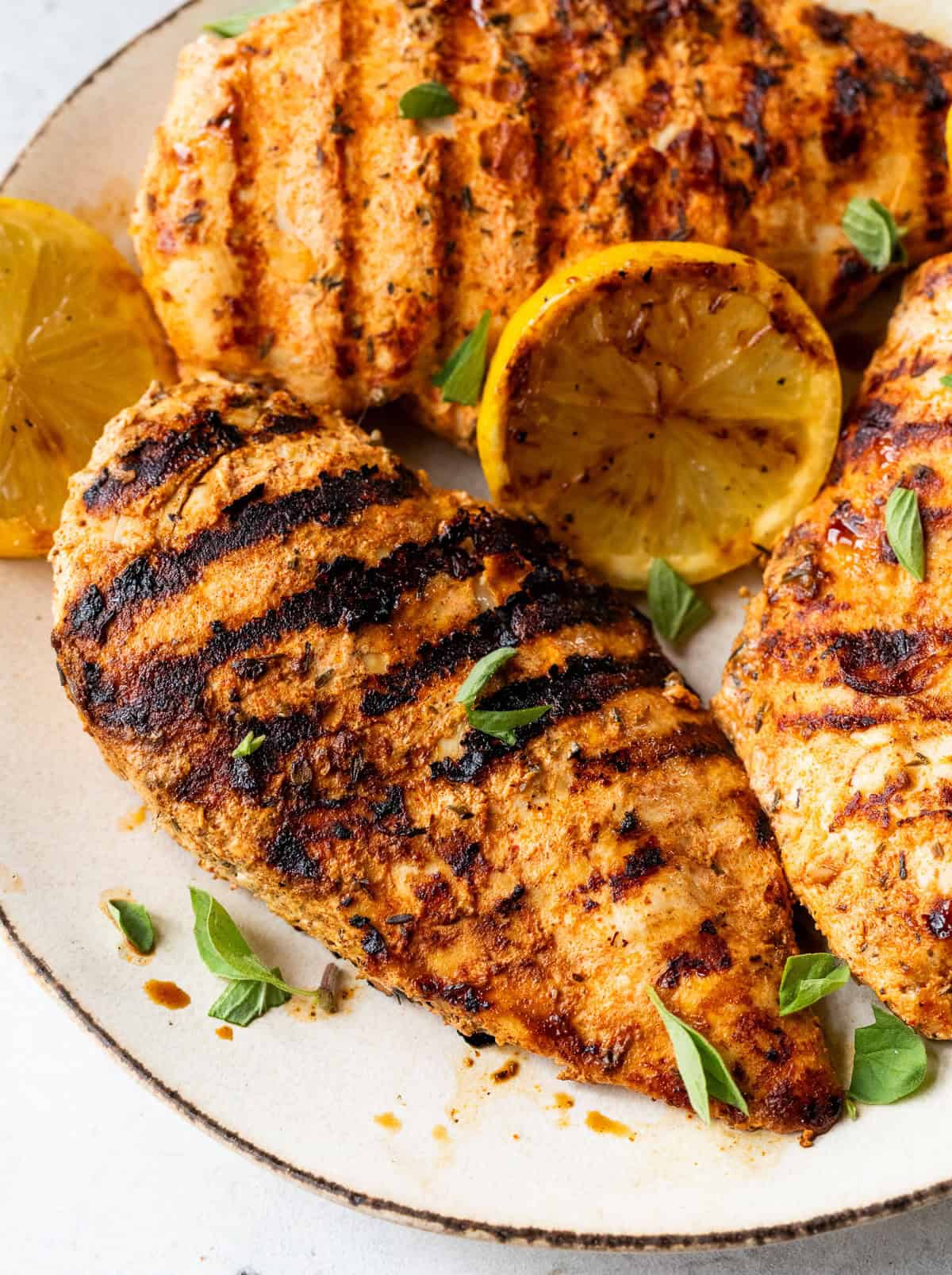 Grilled chicken breasts with grill marks, garnished with fresh herbs and lemon halves on a plate.