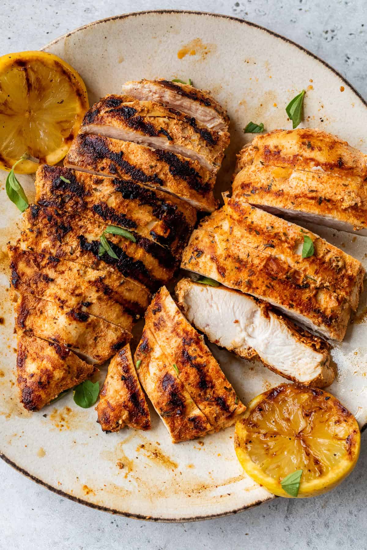 Sliced grilled chicken breast with grill marks, garnished with herbs and served with grilled lemon halves on a plate.