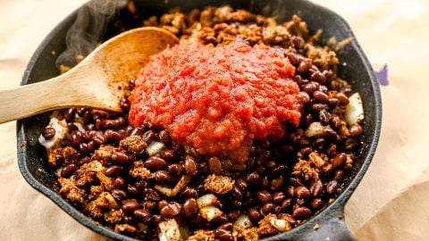 A skillet with beans, ground meat, onions, and tomato sauce, with a wooden spoon resting on the side.
