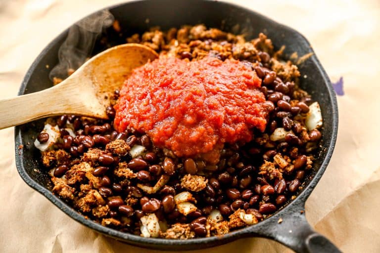 A skillet with beans, ground meat, onions, and tomato sauce, with a wooden spoon resting on the side.