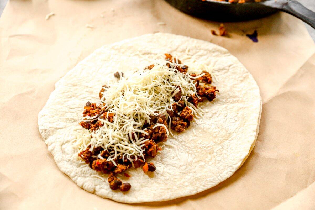 A flour tortilla topped with seasoned ground beef, beans, and shredded cheese on parchment paper.