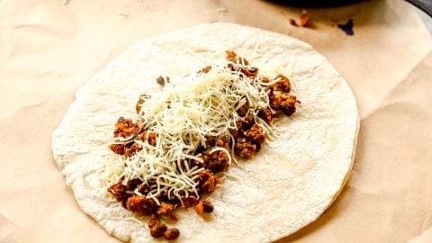 A flour tortilla topped with seasoned ground meat, beans, and shredded cheese on parchment paper.