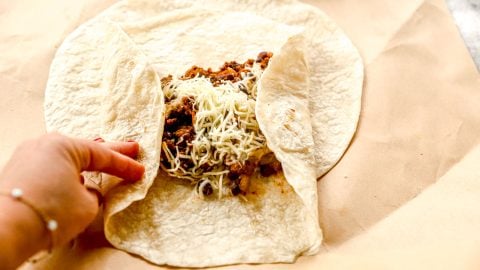 A hand folding a tortilla around seasoned meat and shredded cheese on parchment paper.