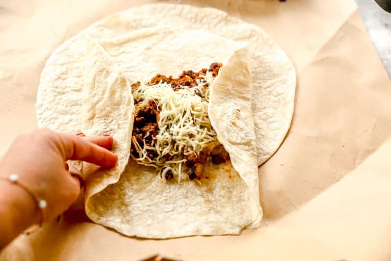 A hand folding a tortilla around seasoned meat and shredded cheese on parchment paper.