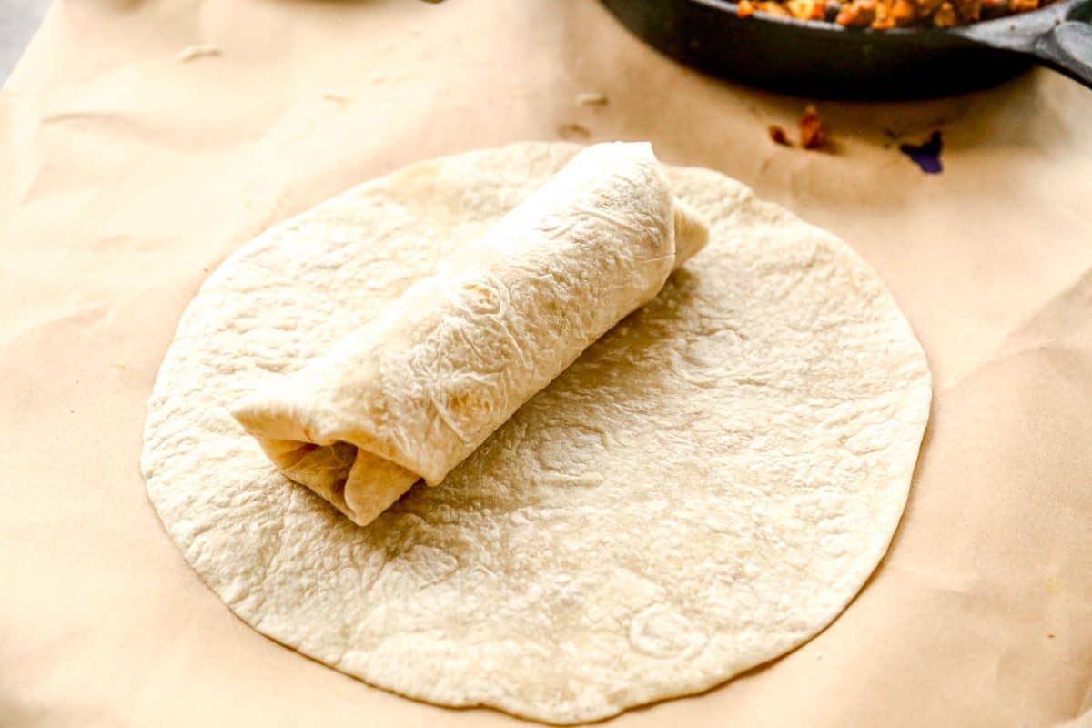 A rolled ground beef burrito sits on top of a flat tortilla with filling visible nearby on parchment paper.