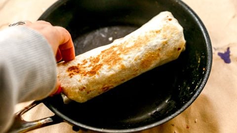 A hand holds a toasted burrito in a black skillet on a light-colored surface.